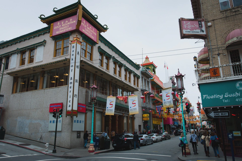 A business called “Gold Mountain Sagely Monastery” on top of a Bank of America with a temple entrance with a turquoise street lamp holding Southwest Airlines ads across the street until the Floating Sushi shop. Both the Floating Sushi and the Bank entrance match the turquoise color of the street lamps whereas the Gold Mountain and the restaurant on top of the Floating Sushi match the red at the top of the street lamps.